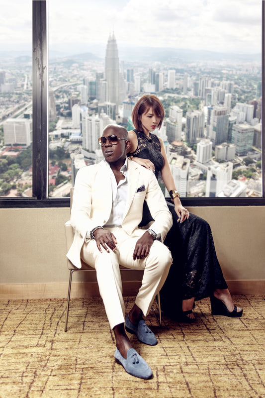 Taiwo Meghoma sitting on a chair wearing tortishell sunglasses, cream two-piece suite with muted blue suede loafers